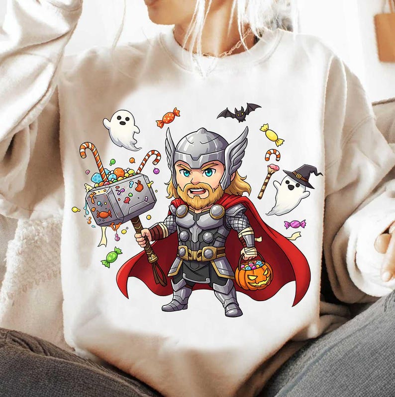 May include: White sweatshirt featuring a cartoon illustration of Thor holding a hammer filled with candy. The design includes Halloween elements like ghosts, bats, and a jack-o'-lantern. The superhero is wearing a helmet, armor, and a red cape.