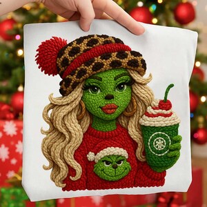 Puede incluir: Un cojín blanco con un personaje verde con cabello rubio, que lleva un suéter rojo y un sombrero con un pompón rojo. El personaje sostiene una taza verde con crema batida y una cereza encima. El cojín tiene un tema navideño.