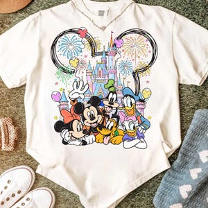 May include: White t-shirt featuring a colorful graphic of Disney characters including Mickey Mouse, Minnie Mouse, Donald Duck, and Goofy, in front of a castle with fireworks and balloons. The shirt has a crew neck and short sleeves.