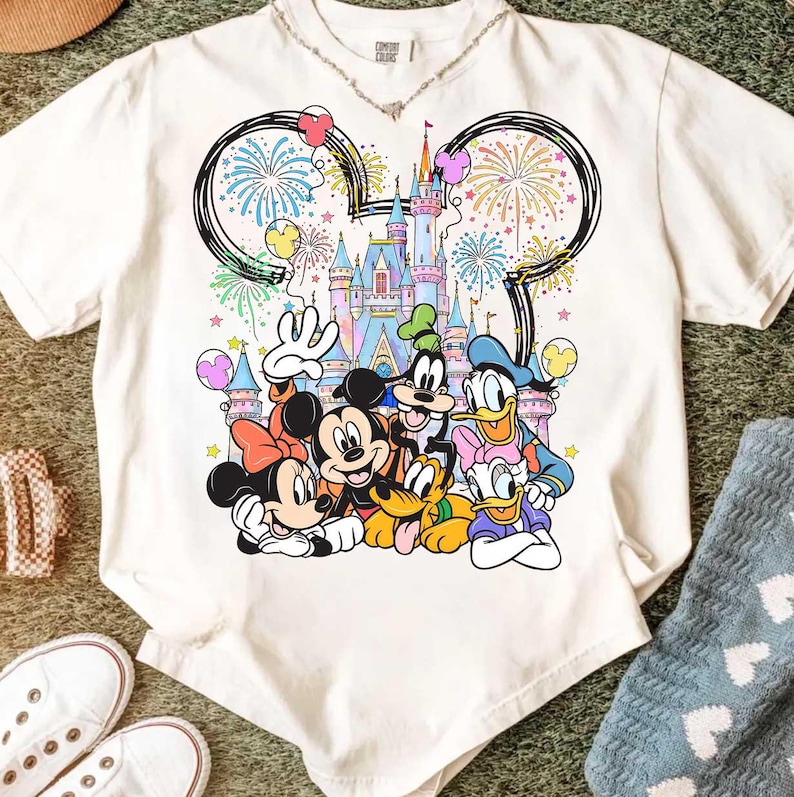 May include: White t-shirt featuring a colorful graphic of Disney characters including Mickey Mouse, Minnie Mouse, Donald Duck, and Goofy, in front of a castle with fireworks and balloons. The shirt has a crew neck and short sleeves.