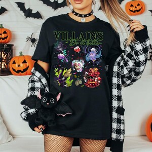 May include: Black t-shirt with the text "Villains Do it better" in green. The shirt features colorful illustrations of various villains. A black bat plush toy is held in the foreground, with Halloween decorations in the background.
