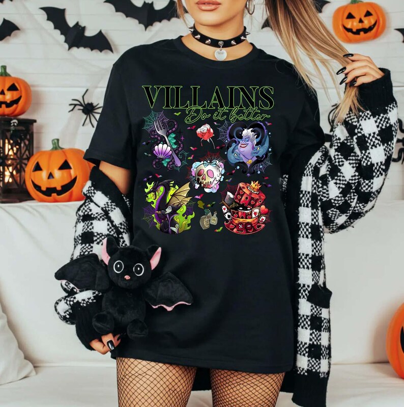 May include: Black t-shirt with the text "Villains Do it better" in green. The shirt features colorful illustrations of various villains. A black bat plush toy is held in the foreground, with Halloween decorations in the background.