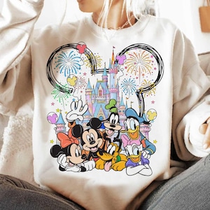 May include: White sweatshirt featuring a colorful graphic of Disney characters, including Mickey and Minnie Mouse, Donald Duck, and Goofy, in front of a castle with fireworks and balloons. The design includes a Mickey Mouse ear outline.
