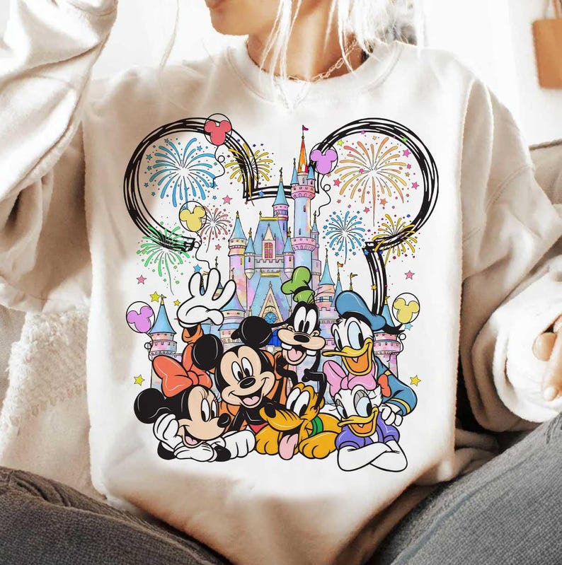 May include: White sweatshirt featuring a colorful graphic of Disney characters, including Mickey and Minnie Mouse, Donald Duck, and Goofy, in front of a castle with fireworks and balloons. The design includes a Mickey Mouse ear outline.