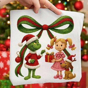 Puede incluir: Un cojín cuadrado blanco con una ilustración navideña. El diseño incluye un personaje verde del Grinch con gorro de Papá Noel, una niña con vestido rosa y un perro con cuernos. Un gran lazo verde y rojo adorna la imagen.