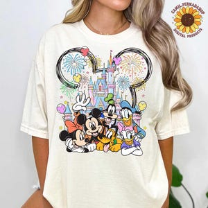 May include: Cream-colored t-shirt featuring a colorful graphic of Disney characters, including Mickey Mouse, Minnie Mouse, Donald Duck, and Goofy, in front of a castle. Fireworks and balloons are also depicted. The shirt has a crew neck.