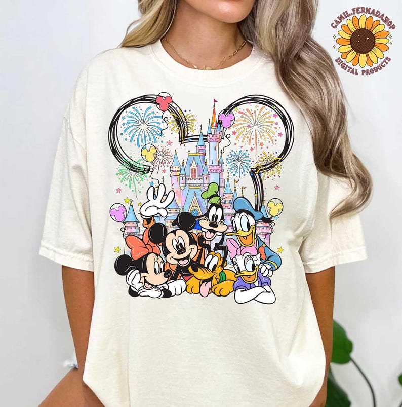 May include: Cream-colored t-shirt featuring a colorful graphic of Disney characters, including Mickey Mouse, Minnie Mouse, Donald Duck, and Goofy, in front of a castle. Fireworks and balloons are also depicted. The shirt has a crew neck.