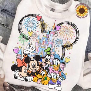 Puede incluir: Sudadera blanca con un gráfico colorido de personajes de Disney, incluyendo Mickey y Minnie Mouse, Goofy, Donald y Daisy Duck, y Pluto. El diseño incluye un castillo, fuegos artificiales y el texto "CAMIL FERNANDEZ DIGITAL PRODUCTS".