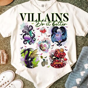 May include: White t-shirt with the text "VILLAINS Do it better" in black and green. The shirt features colorful illustrations of various villains, including a dragon, a skull, and Ursula. The design includes spiderwebs and bats.