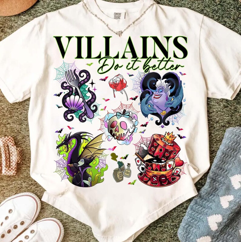 May include: White t-shirt with the text "VILLAINS Do it better" in black and green. The shirt features colorful illustrations of various villains, including a dragon, a skull, and Ursula. The design includes spiderwebs and bats.