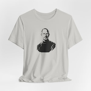 May include: A pale grey t-shirt with a black and white portrait of a man in a black turtleneck. The portrait is centrally placed. The t-shirt has short sleeves and a crew neck.