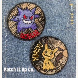 May include: Two embroidered patches with cartoon characters. One shows a purple Gengar with red eyes and the name "GENGAR" in red. The other displays a yellow Mimikyu with the name "MIMIKYU". Both patches have a plaid background.