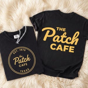 May include: Two black t-shirts with gold lettering. One shirt features "THE Patch CAFE" on the back. The other shirt has a circular design with "EST. 1970 the Patch CAFE TEXAS". A silver chain necklace rests on the shirt.