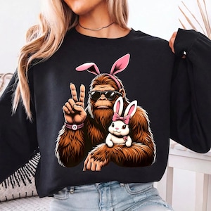 May include: Black sweatshirt featuring a cartoon Bigfoot wearing sunglasses and pink bunny ears, giving a peace sign. A white bunny with a pink bow sits on its arm. The design is a fun, Easter-themed graphic.