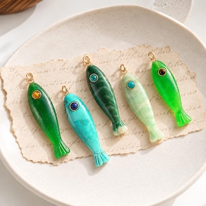 May include: Five fish-shaped pendants in varying shades of green and blue, each featuring a small gold-tone metal loop at the top and a circular gemstone inlay near the head. The pendants are arranged on a piece of textured, off-white paper.