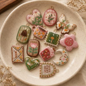 May include: A collection of small, decorative charms in various shapes including hearts, a butterfly, a moon, a mug, and an envelope. Each charm features colorful painted designs such as flowers, mushrooms, and celestial motifs, with some pieces displaying the text SEEDS and COZY VIBES ONLY.