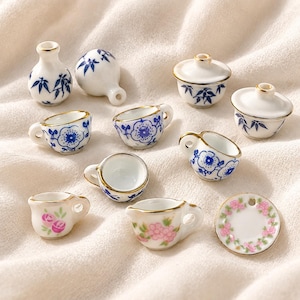 May include: A collection of miniature ceramic items including small cups, lidded bowls, vases, and a plate. Each piece features white porcelain with blue floral or bamboo patterns, or pink rose designs, all accented with gold-painted rims.