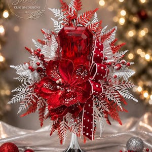 May include: A decorative centerpiece featuring a central red faceted glass pillar candle holder, surrounded by red velvet flowers, silver and red fern-like foliage, and a red ribbon with white pearl accents, all supported by a silver pedestal base.