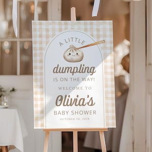 May include: A sign on a wooden easel announces a baby shower. The sign features a dumpling illustration and the text: "A little dumpling is on the way! Welcome to Olivia's Baby Shower October 20, 2028." White balloons decorate the background.