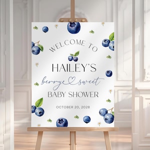 May include: A white baby shower welcome sign on a wooden easel. The sign features watercolor blueberries, leaves, and flowers. Text reads "Welcome to Hailey's berry sweet Baby Shower" with the date October 20, 2028.