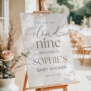 May include: A baby shower welcome sign on a wooden easel. The sign reads "We are on cloud nine, Welcome to Sophie's Baby Shower, August 10, 2028." The background has a soft, cloudy design. A vase of flowers is in the foreground.