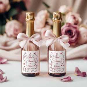 May include: Two small champagne bottles with gold foil tops and pink bows. Each bottle has a white label with the text "That's Amore" and "Olivia's Bridal Shower" in a decorative font. The bottles are set against a backdrop of pink and purple flowers.