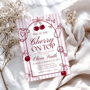 May include: A baby shower invitation with a pink and white striped background. The invitation features a cherry theme with illustrations of cherries, a baby bottle, and a onesie. The text reads "She is the Cherry on Top" and includes event details.