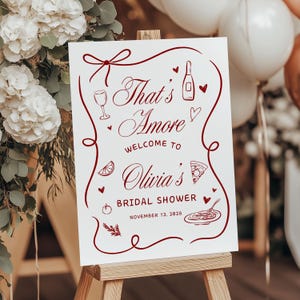 Könnte beinhalten: Ein weißes Schild auf einer hölzernen Staffelei mit dem Text "That's Amore" in burgunderfarbener Schrift, das die Gäste zur Brautparty von Olivia am 12. November 2028 willkommen heißt. Das Schild ist mit Illustrationen von Speisen und Getränken verziert.