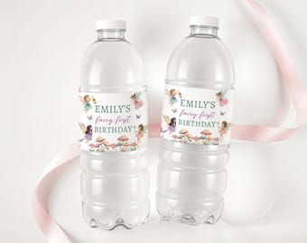 Puede incluir: Dos botellas de agua de plástico transparente con tapas blancas. Cada botella tiene una etiqueta que dice "Emily's fairy first BIRTHDAY" con ilustraciones de hadas, setas y mariposas. Una cinta rosa está en el fondo.