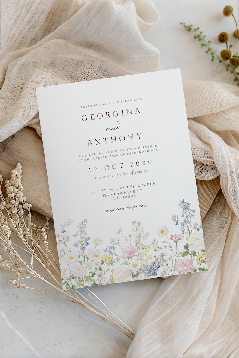 May include: A white rectangular wedding invitation card featuring dark brown serif text and a colourful watercolour floral border at the bottom. The text reads: Together with their families, Georgina and Anthony, request the honour of your presence at the celebration of their marriage, 17 Oct 2030, at 4 o'clock in the afternoon, St. Michael Parish Church, 123 Anywhere St., Any Drive, reception to follow.