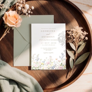 May include: A white wedding invitation card with black text reading Georgina and Anthony, 17 Oct 2030, and St. Michael Parish Church, featuring a colourful floral illustration at the bottom, placed with a sage green envelope and dried flowers.