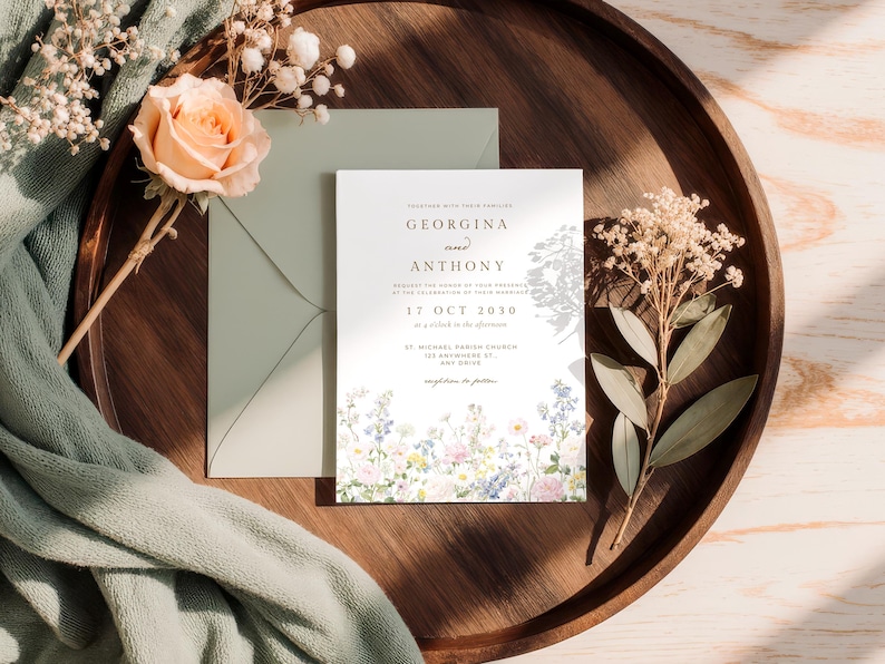 May include: A white wedding invitation card with black text reading Georgina and Anthony, 17 Oct 2030, and St. Michael Parish Church, featuring a colourful floral illustration at the bottom, placed with a sage green envelope and dried flowers.