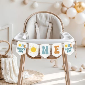 May include: A wooden high chair with a beige seat and a banner that reads "ONE HAPPY BOY." The banner has a yellow sun graphic and is strung across the front of the chair. Balloons in white and gold are in the background.