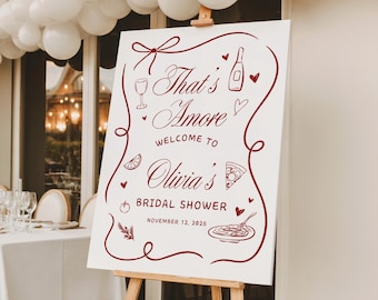 May include: A white sign on a wooden easel with the words "That's Amore" in burgundy script, welcoming guests to Olivia's bridal shower. The sign is decorated with illustrations of a wine glass, bottle, pizza, and other Italian-themed elements.