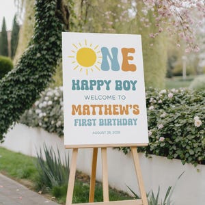 May include: A white sign on a wooden easel with the words "ONE HAPPY BOY" and "WELCOME TO MATTHEW'S FIRST BIRTHDAY AUGUST 28, 2028" in a retro font. The sign has a yellow sun graphic.