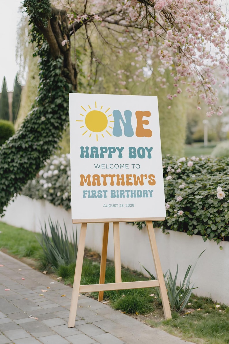 May include: A white sign on a wooden easel with the words "ONE HAPPY BOY" and "WELCOME TO MATTHEW'S FIRST BIRTHDAY AUGUST 28, 2028" in a retro font. The sign has a yellow sun graphic.