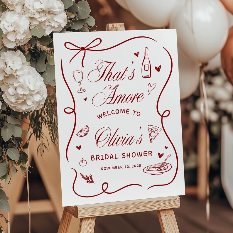 May include: A white sign on a wooden easel with the text "That's Amore" in burgundy script, welcoming guests to Olivia's bridal shower on November 12, 2028. The sign is decorated with burgundy illustrations of food and drink.
