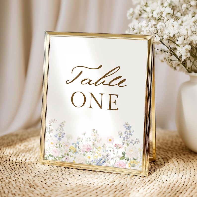 May include: A gold-toned metal frame holding a white card with the text Table ONE in brown script and block lettering. The bottom of the card features a printed illustration of various wildflowers in shades of pink, blue, and yellow.