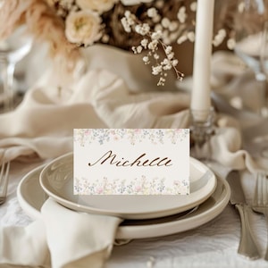 May include: A rectangular place card with a cream-coloured background featuring a delicate floral border of small pink, blue, and yellow flowers at the top and bottom edges. The name Michelle is printed in the centre in a brown cursive script font.