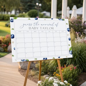May include: A white sign on a wooden easel with the text "guess the arrival of BABY TAYLOR" and a calendar grid. The sign is decorated with blueberry and leaf illustrations. The easel is set up outdoors near flowers and greenery.