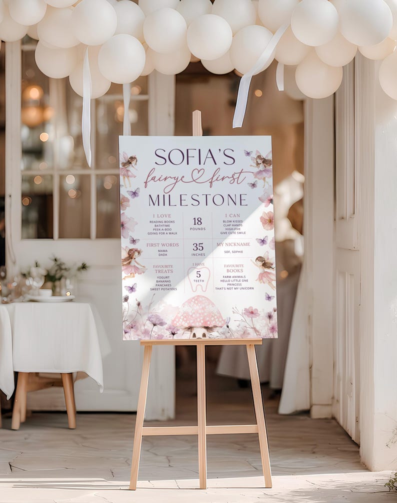 Puede incluir: Un caballete de madera exhibe un letrero de hito para "Sofia's Fairye First Milestone". El letrero presenta un tema de hadas con detalles florales y mariposas, e incluye detalles como "8 kg" y "89 cm."