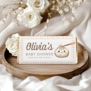 May include: A chocolate bar with a light brown and white checkered pattern. The label reads "Olivia's BABY SHOWER THANK YOU FOR COMING!" with a dumpling illustration. The bar is on a wooden tray with white flowers in the background.