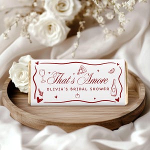 May include: A white candy bar wrapper with the text "That's Amore" in a burgundy script, along with illustrations of a wine glass, pizza slice, and other food-related icons. The text "Olivia's Bridal Shower" is also included.