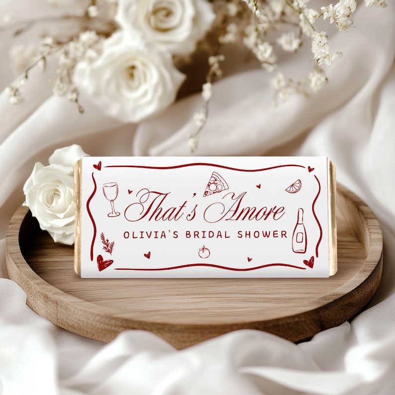 May include: A white candy bar wrapper with the text "That's Amore" in a burgundy script, along with illustrations of a wine glass, pizza slice, and other food-related icons. The text "Olivia's Bridal Shower" is also included.