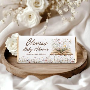 Puede incluir: Envoltorio rectangular de barra de chocolate con el texto "Olivia's Baby Shower" en escritura marrón. El envoltorio presenta una ilustración de un libro abierto lleno de flores y un borde floral. También se incluye el texto "Thank you for coming!"