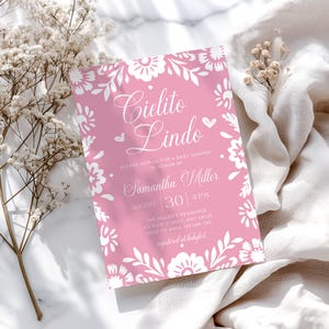 May include: A rectangular baby shower invitation card with a pink background and white floral border. The text reads Cielito Lindo, Please join us for a baby shower in honor of Samantha Miller, August 30, 4 PM, The Miller's Residence, 123 Main Street, Any Drive, RSVP to Anne 123 456 789, registered at Babylist.
