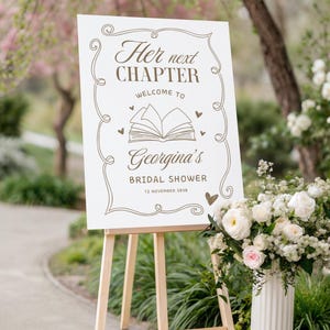 May include: A white sign on a wooden easel with the text "Her next CHAPTER Welcome to Georgina's BRIDAL SHOWER 12 NOVEMBER 2030" in brown script. The sign is decorated with hearts and an open book illustration. A vase of white and pink flowers is to the right.