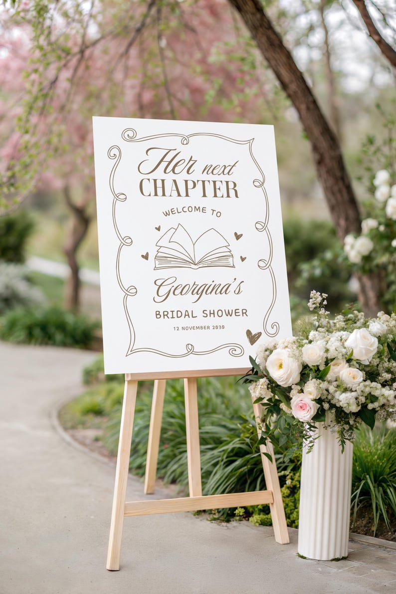 May include: A white sign on a wooden easel with the text "Her next CHAPTER Welcome to Georgina's BRIDAL SHOWER 12 NOVEMBER 2030" in brown script. The sign is decorated with hearts and an open book illustration. A vase of white and pink flowers is to the right.