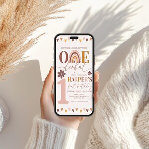 Peut inclure: Un smartphone affiche une invitation d'anniversaire avec le texte "ONE derful" et "Harper's first birthday". Le design comprend un arc-en-ciel, des fleurs et des c&oelig;urs dans des tons de rose, de marron et d'or. Le t&eacute;l&eacute;phone est tenu sur un fond doux et neutre.