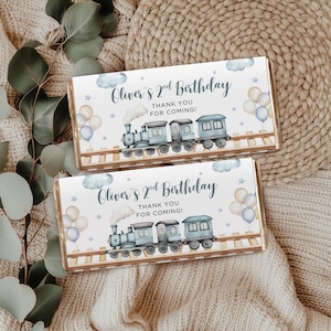 Op de afbeelding: Twee chocoladereepverpakkingen met een treinillustratie. De verpakkingen lezen "Oliver's 2nd Birthday" en "Thank You For Coming!". De trein is lichtblauw, met ballonnen en wolken op de achtergrond.